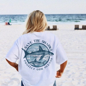 May include: White t-shirt with a blue circle graphic on the back. The graphic features three sharks swimming in waves and the text "Save the Sharks Protect Our Oceans".