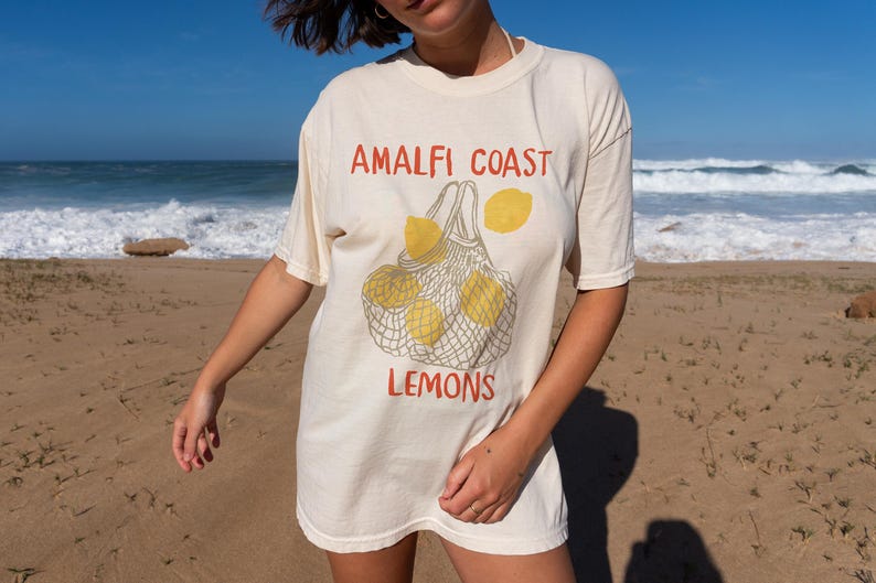 May include: A white t-shirt with a graphic of lemons in a mesh bag. The text "Amalfi Coast Lemons" is printed on the shirt.