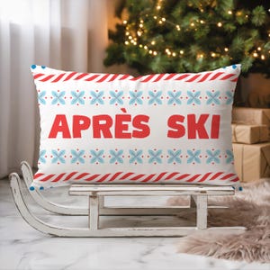 Può includere: Cuscino rettangolare bianco con dettagli rossi e blu, con la scritta "APRÈS SKI" in rosso. Il design include strisce rosse e bianche e motivi floreali blu. Il cuscino è appoggiato su una slitta di legno bianca.