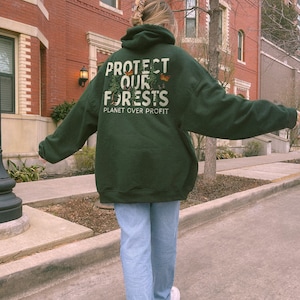 May include: A dark green hooded sweatshirt with the text "Protect Our Forests" and "Planet Over Profit" printed on the back. The sweatshirt is worn by a person walking down a sidewalk.