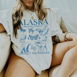 Puede incluir: Camiseta blanca con un estampado gr&aacute;fico azul del texto "ALASKA the last frontier" y varios animales salvajes de Alaska, incluyendo una ballena, una mariposa, un oso, un alce y un ciervo. El texto "PROTECT THE WILDLIFE" est&aacute; impreso debajo del gr&aacute;fico.