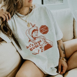 May include: A white t-shirt with a red graphic of a woman in a cowboy hat reading a book. The graphic says "let's read girls!" and "saddle up with a good book".