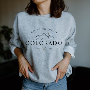 K&ouml;nnte beinhalten: Hellgraues Sweatshirt mit dem Text "Aspen Ski Resort Colorado est. 1879" in Schwarz und Gold. Das Design zeigt eine Bergkette und gekreuzte Skist&ouml;cke. Das Sweatshirt ist ein l&auml;ssiger Stil.