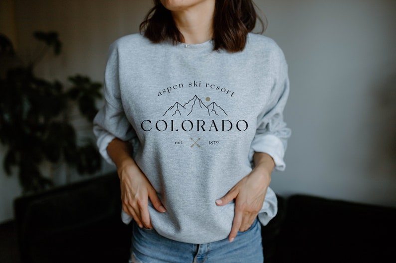 K&ouml;nnte beinhalten: Hellgraues Sweatshirt mit dem Text "Aspen Ski Resort Colorado est. 1879" in Schwarz und Gold. Das Design zeigt eine Bergkette und gekreuzte Skist&ouml;cke. Das Sweatshirt ist ein l&auml;ssiger Stil.
