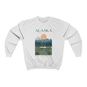 Pu&ograve; includere: Felpa bianca con un design grafico di una catena montuosa con un tramonto e il testo "ALASKA LAND OF THE MIDNIGHT SUN".