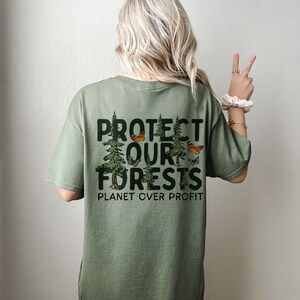 Op de afbeelding: Groen T-shirt met een grafische print van bomen en vlinders. De tekst "PROTECT OUR FORESTS PLANET OVER PROFIT" staat op de achterkant van het shirt.