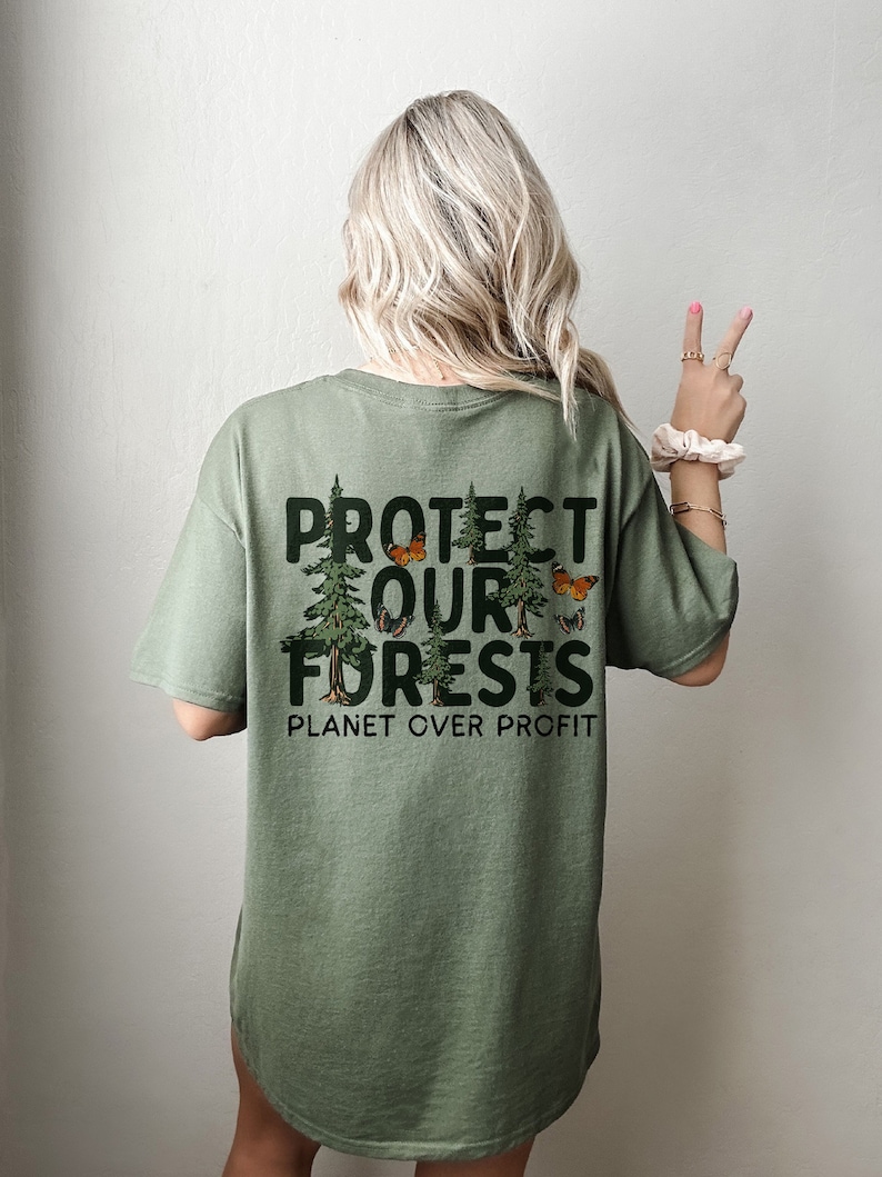 Op de afbeelding: Groen T-shirt met een grafische print van bomen en vlinders. De tekst "PROTECT OUR FORESTS PLANET OVER PROFIT" staat op de achterkant van het shirt.