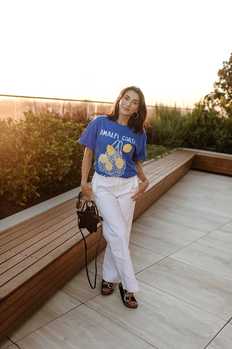 May include: A woman wearing a blue t-shirt with the text "Amalfi Coast" and a lemon graphic, white pants, and black sandals. She is standing on a patio with a wooden bench behind her.