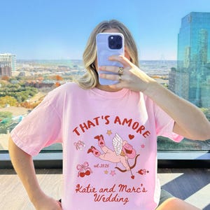 K&ouml;nnte beinhalten: Rosa T-Shirt mit einem Grafikdruck eines Cupidos mit Pfeil und Bogen, Herzen und Sternen. Der Text "That's Amore" befindet sich &uuml;ber dem Grafikdruck und "Katie and Marc's Wedding" darunter. Der Text "est. 2025" befindet sich in der Mitte des Grafikdrucks.