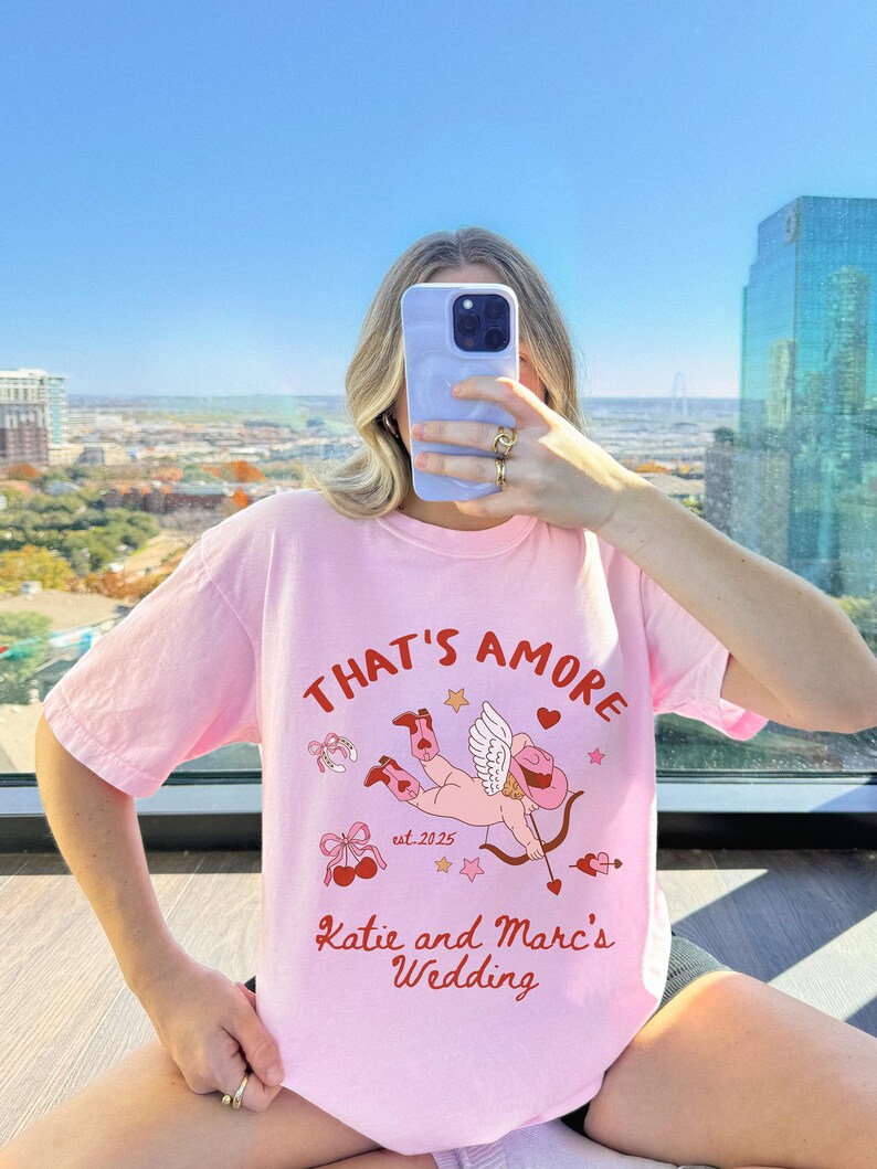K&ouml;nnte beinhalten: Rosa T-Shirt mit einem Grafikdruck eines Cupidos mit Pfeil und Bogen, Herzen und Sternen. Der Text "That's Amore" befindet sich &uuml;ber dem Grafikdruck und "Katie and Marc's Wedding" darunter. Der Text "est. 2025" befindet sich in der Mitte des Grafikdrucks.