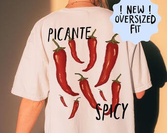 Op de afbeelding: Wit oversized T-shirt met een rode chilipeper print. Het shirt heeft de zwarte woorden "PICANTE" en "SPICY", en de tekst "! NEW ! OVERSIZED FIT" in een blauwe tekstballon. Het ontwerp bevat meerdere rode chilipepers.