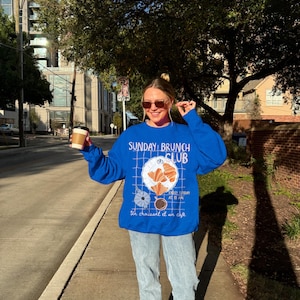 May include: A woman wearing a blue sweatshirt with the text "SUNDAY BRUNCH CLUB" and a graphic of a croissant and coffee. The text below the graphic reads "Every Sunday at 11 AM. Un croissant et un cafe."
