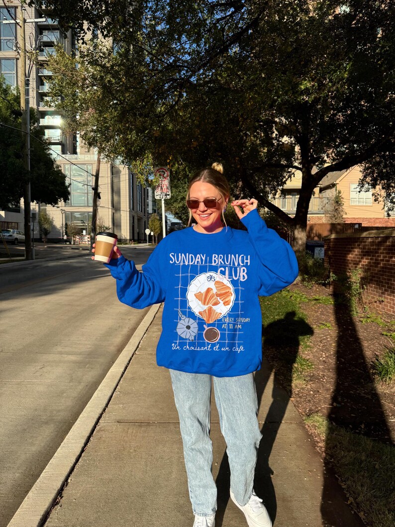 May include: A woman wearing a blue sweatshirt with the text "SUNDAY BRUNCH CLUB" and a graphic of a croissant and coffee. The text below the graphic reads "Every Sunday at 11 AM. Un croissant et un cafe."