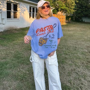 May include: A woman wearing a light blue t-shirt with the text "PASTA Lover Club" and a graphic of a fork with spaghetti. The shirt also says "consumed daily 7 pm - 10 pm". She is wearing white trousers and a white baseball cap.