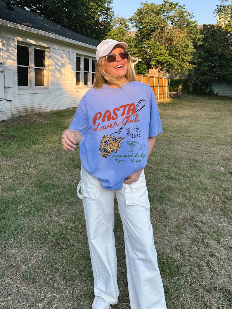May include: A woman wearing a light blue t-shirt with the text "PASTA Lover Club" and a graphic of a fork with spaghetti. The shirt also says "consumed daily 7 pm - 10 pm". She is wearing white trousers and a white baseball cap.