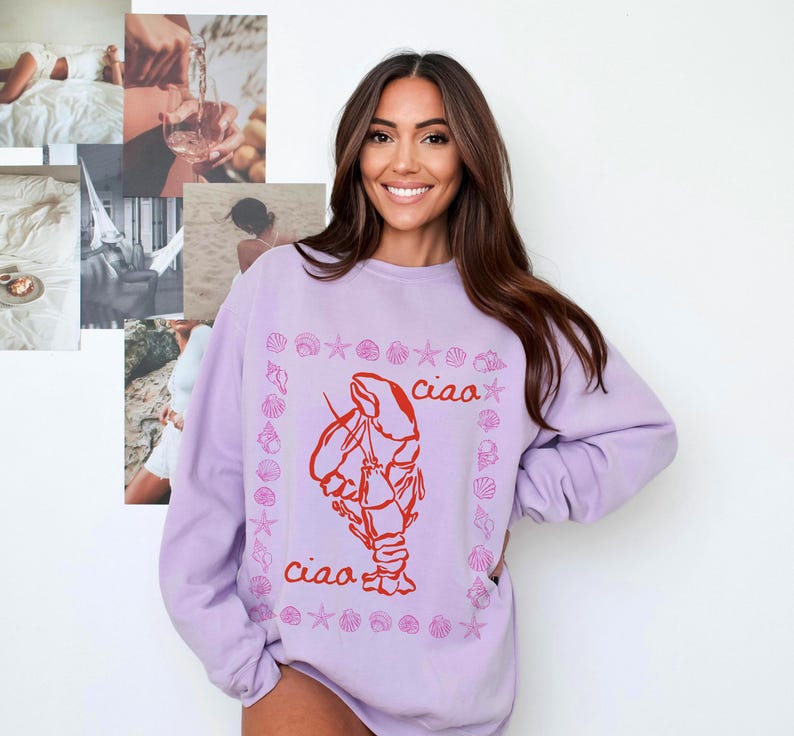 Pu&ograve; includere: Una felpa color lavanda con una grafica di aragosta rossa e il testo "ciao" ripetuto due volte. L'aragosta &egrave; circondata da un bordo di conchiglie rosa.