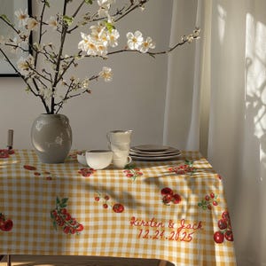 May include: A yellow and white checkered tablecloth with a red tomato and green vine design. The tablecloth has the text "Kirsten & Jack 12.24.2025" printed in red.