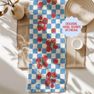 May include: A blue and white checkered table runner with a repeating pattern of red tomatoes. The runner is made of fabric and has the text "ORIGINAL HAND DRAWN ARTWORK" on it. The runner is displayed on a beige surface.