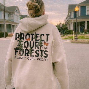 May include: A beige hoodie with a graphic print of trees and butterflies. The text on the hoodie reads "Protect Our Forests Planet Over Profit".
