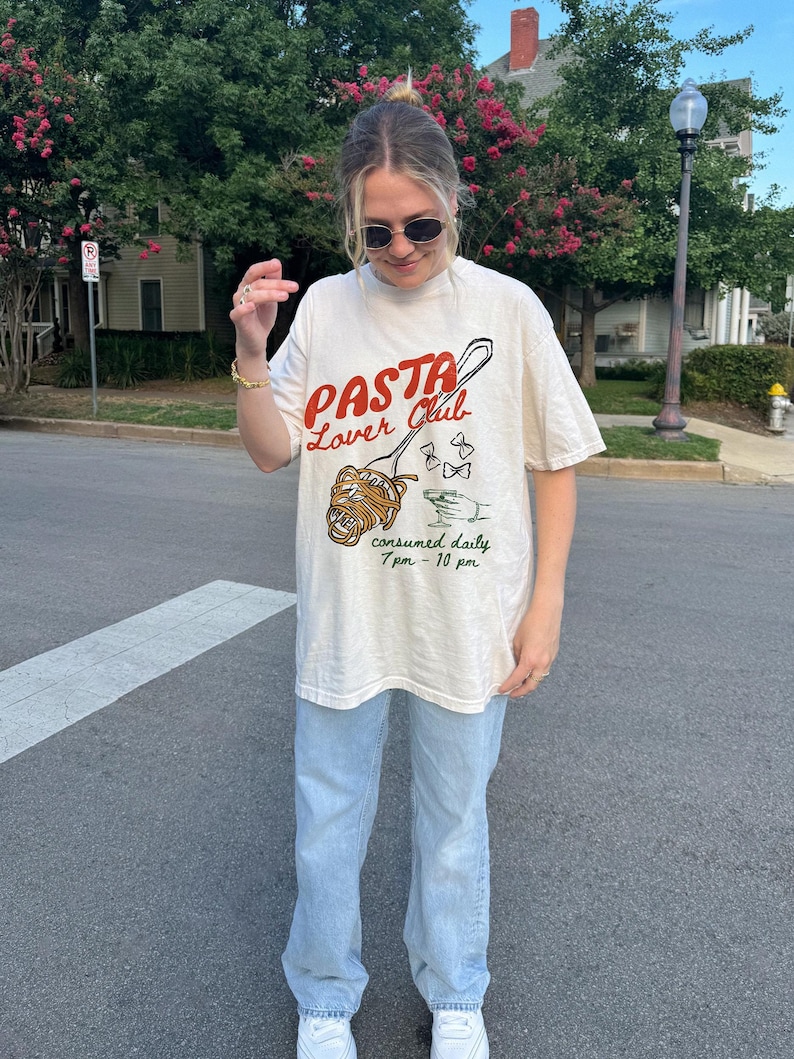 May include: A white t-shirt with a graphic of a pasta dish on a spoon with the text "Pasta Lover Club" and "consumed daily 7pm - 10pm".