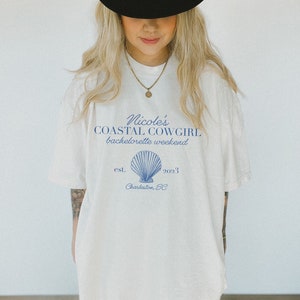 K&ouml;nnte beinhalten: Wei&szlig;es T-Shirt mit einem blauen Grafikdesign, auf dem "Nicole's Coastal Cowgirl bachelorette weekend est. 2023 Charleston, SC" steht, mit einer blauen Muschelillustration.