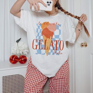 K&ouml;nnte beinhalten: Wei&szlig;es T-Shirt mit einem Aufdruck von drei Kugeln Eis in einer Waffeltüte auf einem blau-wei&szlig; karierten Hintergrund. Der Text "la dolce vita GELATO Italia" ist auf dem Shirt gedruckt.