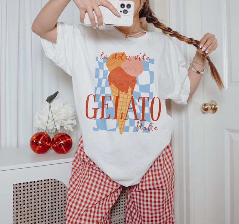 K&ouml;nnte beinhalten: Wei&szlig;es T-Shirt mit einem Aufdruck von drei Kugeln Eis in einer Waffeltüte auf einem blau-wei&szlig; karierten Hintergrund. Der Text "la dolce vita GELATO Italia" ist auf dem Shirt gedruckt.