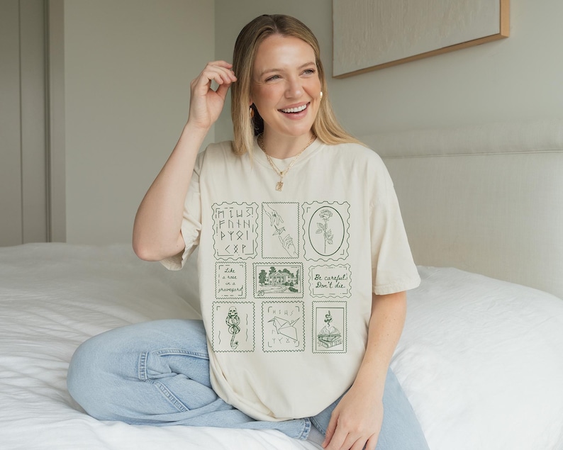 May include: Cream-colored t-shirt featuring a collection of green postage stamp designs. The stamps depict various illustrations, including a hand, a flower, and a snake. The shirt also includes the text "Be careful, don't die."