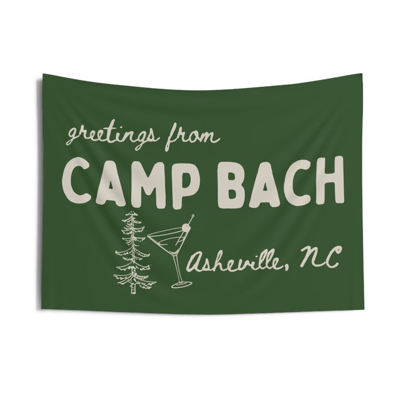 Op de afbeelding: Een groene vlag met witte tekst die "greetings from CAMP BACH Asheville, NC" luidt. Op de vlag zijn ook een kleine dennenboom en een martini-glas met een olijf afgebeeld.