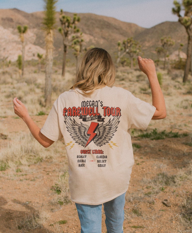 May include: A beige t-shirt with a graphic of wings and a lightning bolt. The text "Mecan's Farewell Tour May 2024" is printed on the shirt. The text "Guest Stars" is printed below the graphic, followed by a list of names.