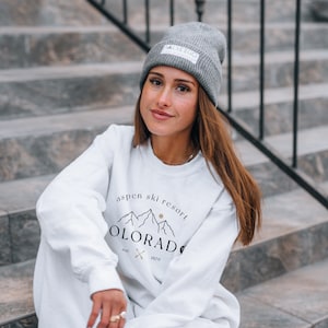 K&ouml;nnte beinhalten: Eine graue M&uuml;tze mit wei&szlig;em Etikett und dem Text "ALSEEGO". Ein wei&szlig;es Sweatshirt und eine passende Jogginghose mit dem Text "Aspen Ski Resort Colorado" und einer Berg-Grafik. Das Outfit wird von einer Person getragen, die auf grauen Stufen sitzt.