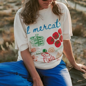 May include: A woman wearing a white t-shirt with a colorful illustration of food and the text "il mercato". She is also wearing a brown baseball cap and blue pants.