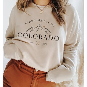 K&ouml;nnte beinhalten: Beiges Sweatshirt mit dem Text "Aspen Ski Resort Colorado" in Schwarz. Das Design umfasst Berggrafiken, eine Sonne und gekreuzte Skist&ouml;cke. Das Sweatshirt wird mit einer braunen Hose kombiniert.
