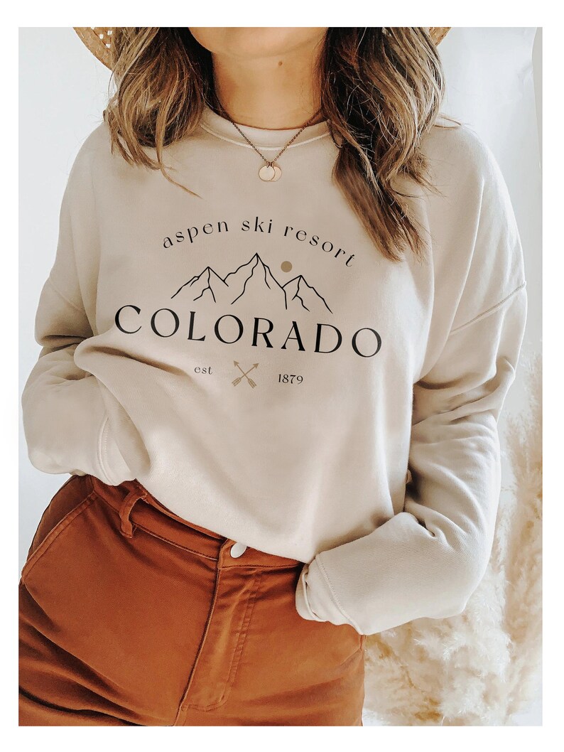 K&ouml;nnte beinhalten: Beiges Sweatshirt mit dem Text "Aspen Ski Resort Colorado" in Schwarz. Das Design umfasst Berggrafiken, eine Sonne und gekreuzte Skist&ouml;cke. Das Sweatshirt wird mit einer braunen Hose kombiniert.