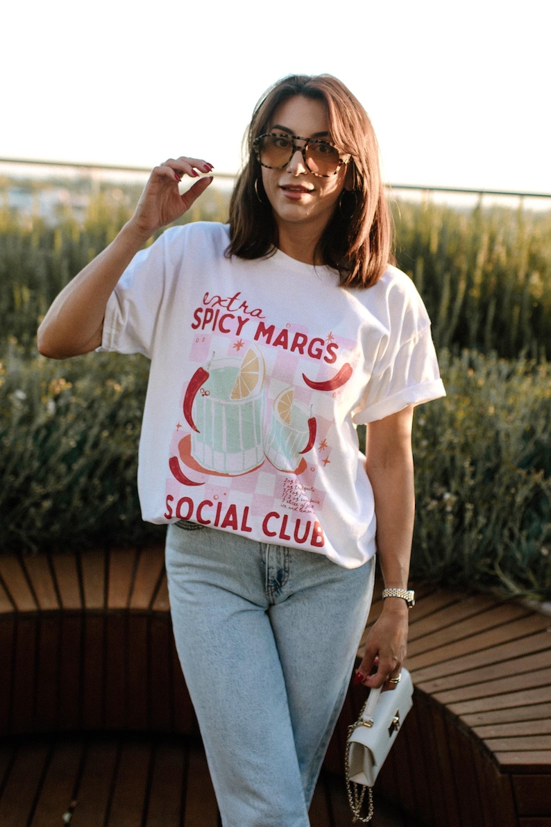 May include: A white t-shirt with a graphic of a cocktail drink and the text "extra spicy margs social club". The person wearing the shirt is also wearing blue jeans and sunglasses.