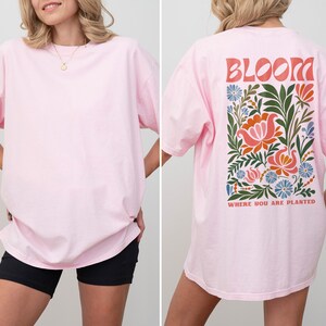 Puede incluir: Camiseta rosa claro con un dise&ntilde;o floral en la espalda. El dise&ntilde;o incluye la palabra "BLOOM" en letras naranjas sobre una disposici&oacute;n rectangular de flores en tonos rosa, naranja, azul y verde. El texto "WHERE YOU ARE PLANTED" est&aacute; debajo.