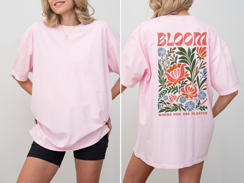 Puede incluir: Camiseta rosa claro con un dise&ntilde;o floral en la espalda. El dise&ntilde;o incluye la palabra "BLOOM" en letras naranjas sobre una disposici&oacute;n rectangular de flores en tonos rosa, naranja, azul y verde. El texto "WHERE YOU ARE PLANTED" est&aacute; debajo.