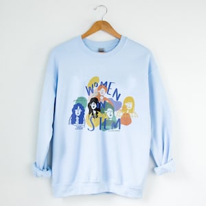May include: Light blue sweatshirt with a graphic print of women in STEM. The graphic features colorful illustrations of women's faces and the text "Women in STEM".