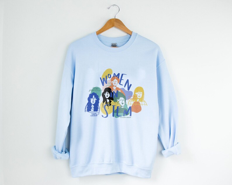 May include: Light blue sweatshirt with a graphic print of women in STEM. The graphic features colorful illustrations of women's faces and the text "Women in STEM".