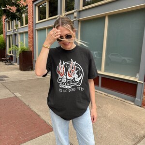 May include: A black t-shirt with a skull graphic and flames coming out of the eyes. The text "Is he dead yet?" is printed below the skull. The person wearing the shirt is standing on a sidewalk in front of a brick building.