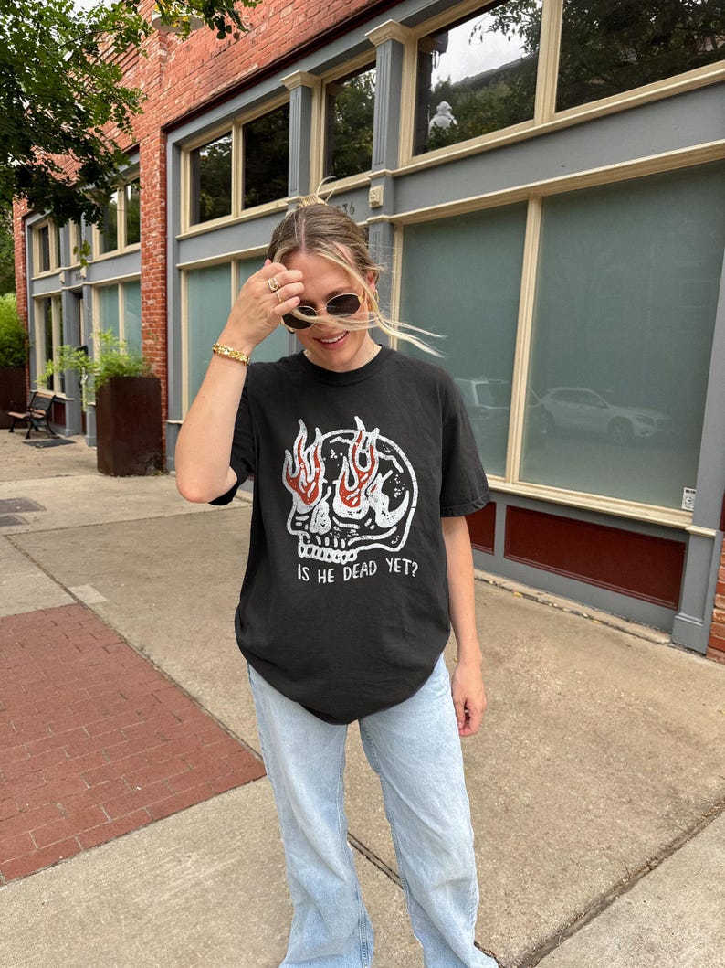 May include: A black t-shirt with a skull graphic and flames coming out of the eyes. The text "Is he dead yet?" is printed below the skull. The person wearing the shirt is standing on a sidewalk in front of a brick building.