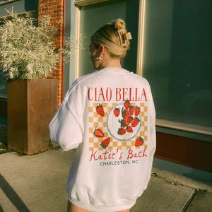Peut inclure: Sweat-shirt blanc avec un motif rouge et jaune de fraises sur une assiette avec le texte "Ciao Bella" et "Katie's Bach Charleston, NC" imprim&eacute; au dos.