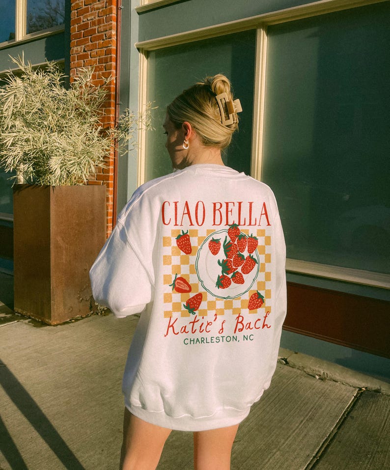 Peut inclure: Sweat-shirt blanc avec un motif rouge et jaune de fraises sur une assiette avec le texte "Ciao Bella" et "Katie's Bach Charleston, NC" imprim&eacute; au dos.