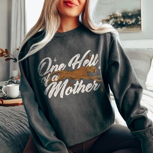 Peut inclure: Un sweat-shirt gris fonc&eacute; avec un imprim&eacute; graphique de style vintage qui dit "One Hell of a Mother" avec un gu&eacute;pard courant sur le texte.