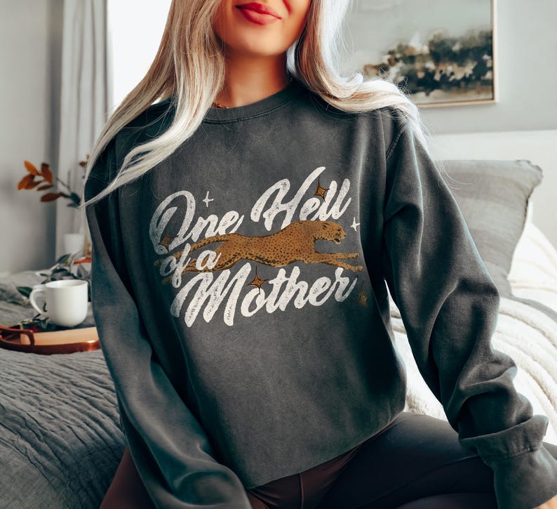 Peut inclure: Un sweat-shirt gris fonc&eacute; avec un imprim&eacute; graphique de style vintage qui dit "One Hell of a Mother" avec un gu&eacute;pard courant sur le texte.