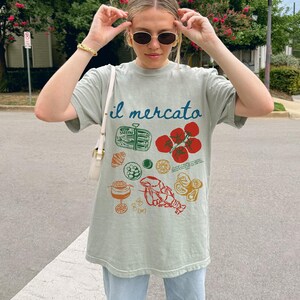 Op de afbeelding: Een lichtgroen T-shirt met de tekst "il mercato" erop gedrukt. Het shirt heeft een grafisch ontwerp met verschillende Italiaanse voedingsmiddelen, waaronder tomaten, pasta, zeevruchten en een glas wijn.