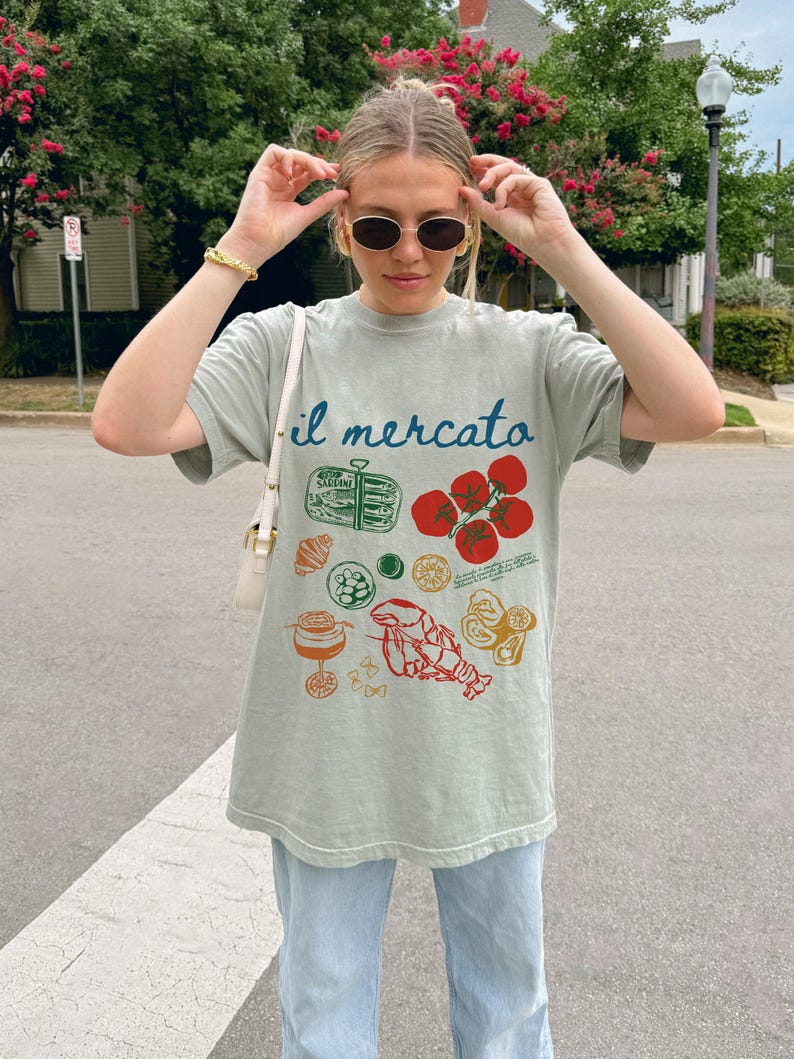 Op de afbeelding: Een lichtgroen T-shirt met de tekst "il mercato" erop gedrukt. Het shirt heeft een grafisch ontwerp met verschillende Italiaanse voedingsmiddelen, waaronder tomaten, pasta, zeevruchten en een glas wijn.