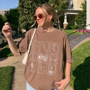 May include: Brown t-shirt with a stamp-like design featuring various illustrations and the text "Be careful, Don't die." The shirt is paired with light blue jeans. The person is wearing sunglasses and a white purse.