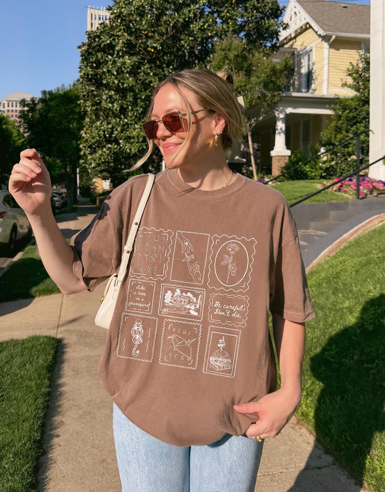 May include: Brown t-shirt with a stamp-like design featuring various illustrations and the text "Be careful, Don't die." The shirt is paired with light blue jeans. The person is wearing sunglasses and a white purse.