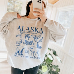 May include: Grey sweatshirt with a blue graphic print of the word "Alaska" and the text "the last frontier". The graphic also includes images of animals such as a bear, moose, whale, butterfly, and birds. The text "Protect the Wildlife" is printed below the graphic.
